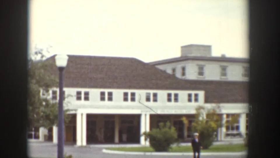 Wyoming 1939 home-movie frame showing a white institutional-looking building, brown roof, porch columns, grounds, and a distant walker.