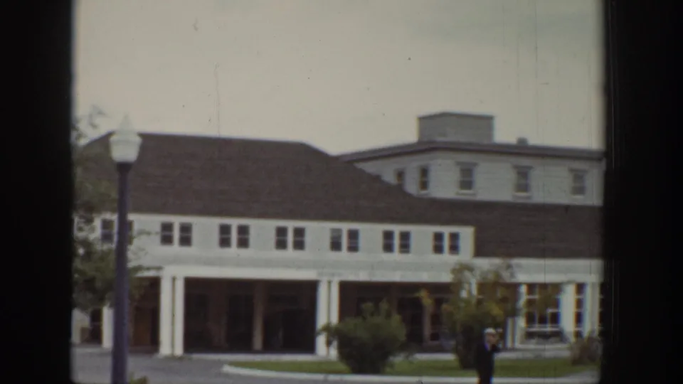 Wyoming 1939 frame of a white building exterior with brown roof, lampposts, landscaped walkway, and a distant person near the front.