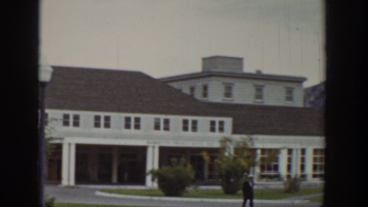 1939 Wyoming home-movie frame showing a white porch-front building, a person walking nearby, and rolling hillside beyond the town street.