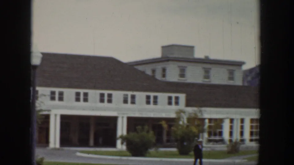 1939 Wyoming home-movie frame showing a white porch-front building, a person walking nearby, and rolling hillside beyond the town street.