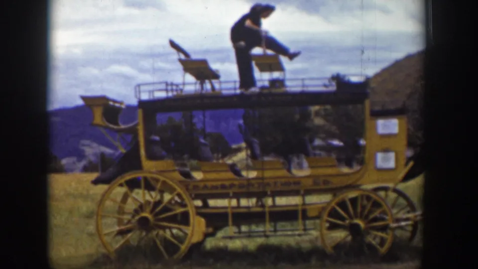 In the Wyoming 1939 clip, a man bends forward on the roof platform of a yellow stagecoach-style vehicle, with side lettering visible below.