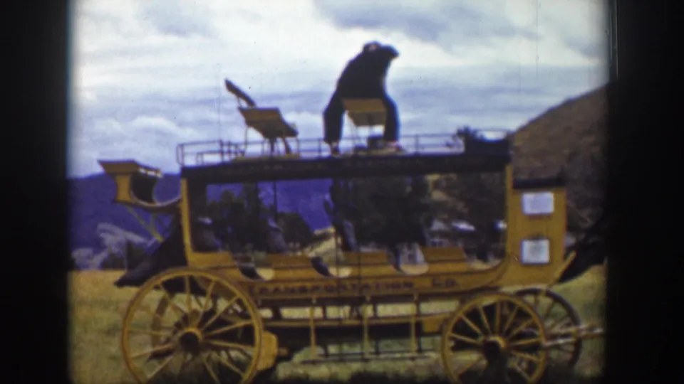 A man crouches near a roof seat on a yellow stagecoach-style vehicle in 1939 Wyoming footage, with hills and cloudy sky behind him.