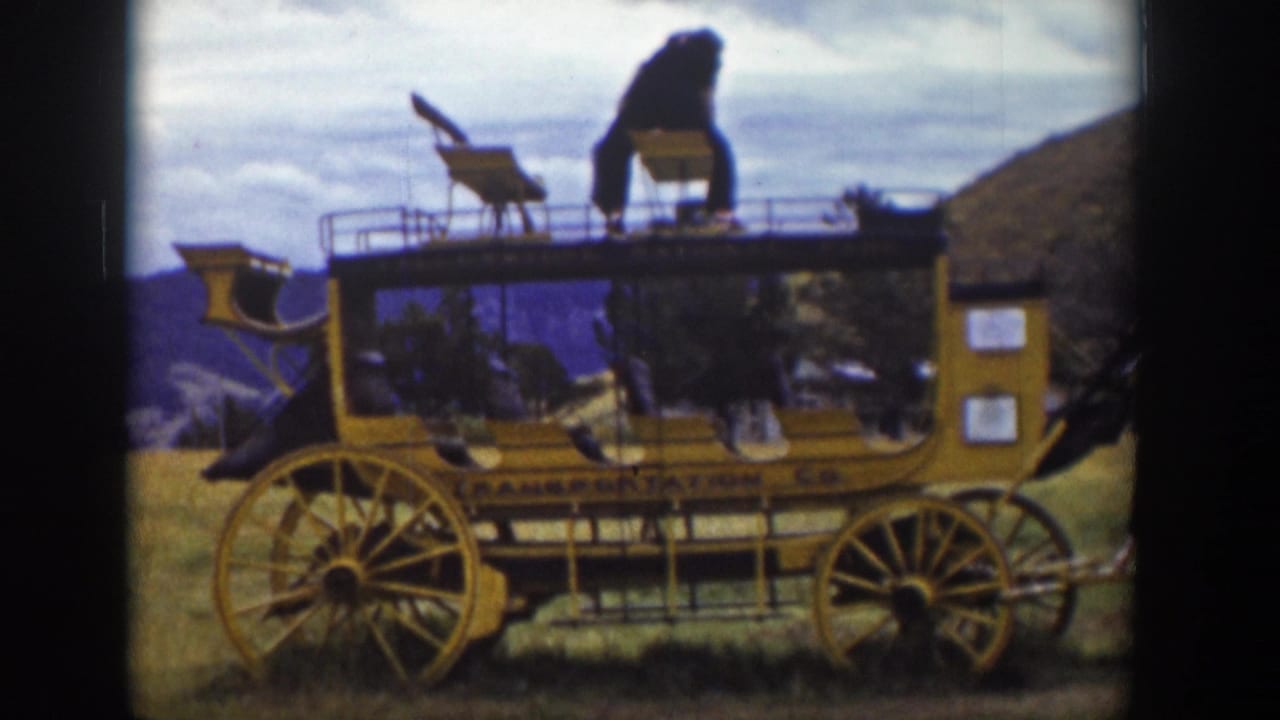 1939 Wyoming 8mm frame of a yellow stagecoach with large wooden wheels and a person standing on the roof against hills and cloudy sky.