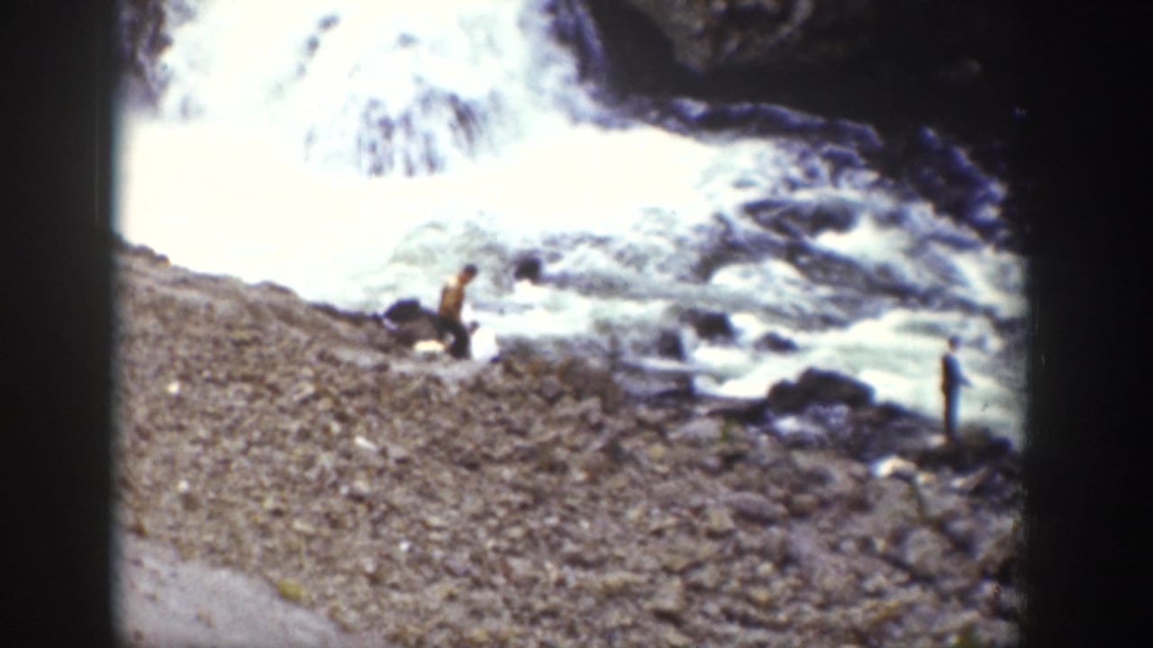 1939 Wyoming 8mm frame showing distant people near a rocky riverbank as white waterfall water rushes behind them.