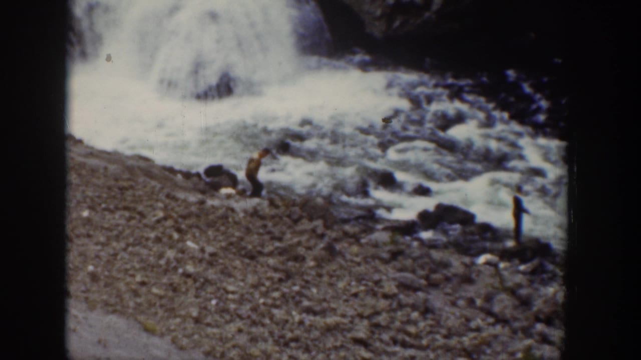 1939 Wyoming home-movie frame with distant visitors beside a rocky waterfall edge and foaming water.