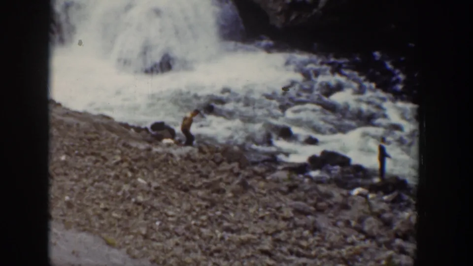 1939 Wyoming home-movie frame with distant visitors beside a rocky waterfall edge and foaming water.