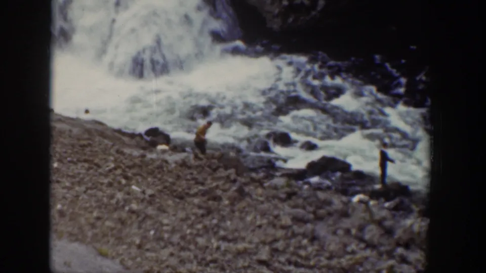 1939 Wyoming 8mm frame showing distant people on wet rocks near a rushing waterfall and gravel bank.
