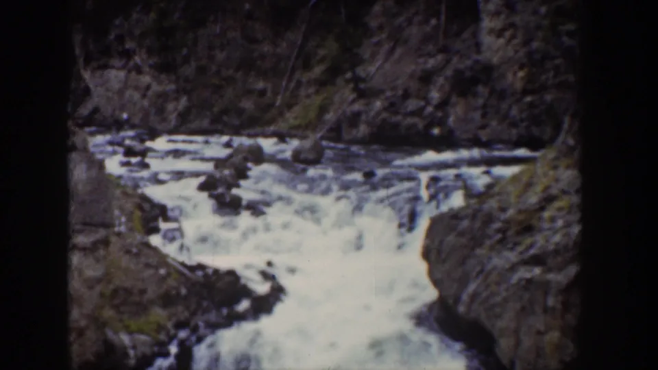 1939 Wyoming home-movie frame showing foaming water moving through rocks beneath a wooded slope, with no clear figure visible.