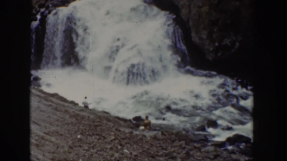 1939 Wyoming home-movie frame showing the waterfall base, rocky shore, and small visitors near the rushing water.