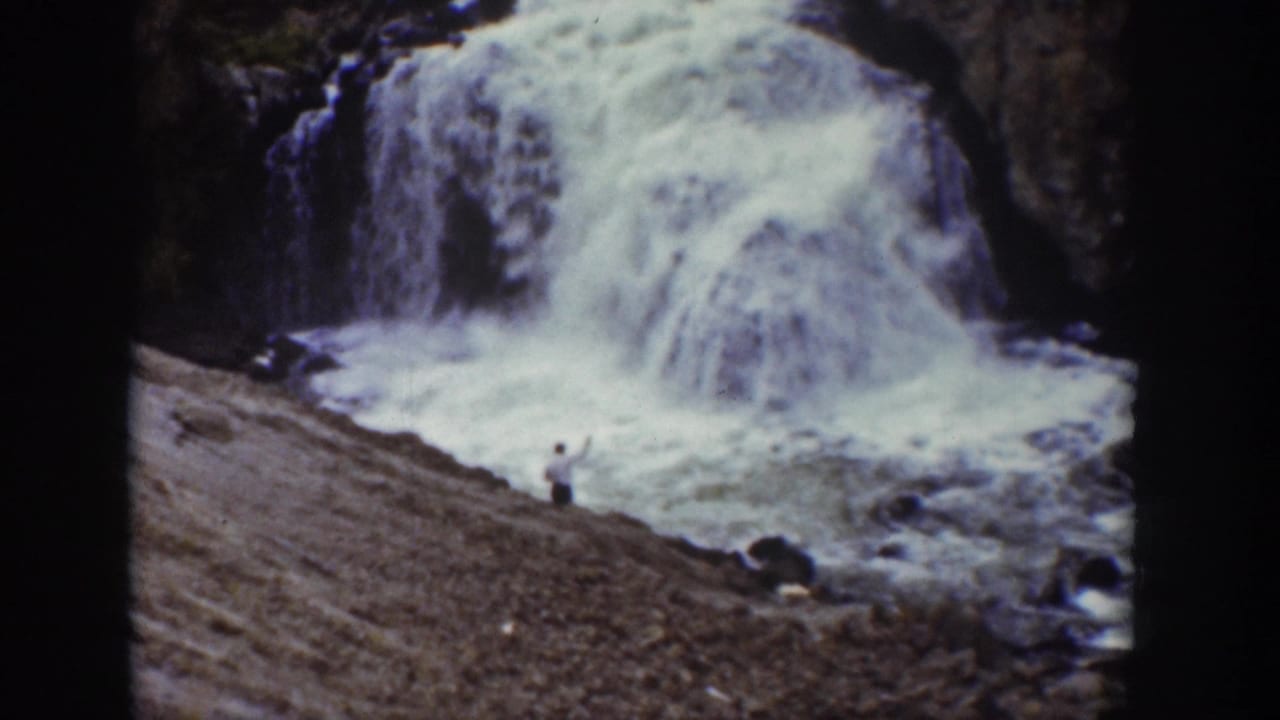Man on a rocky slope facing a broad waterfall and foaming water.
