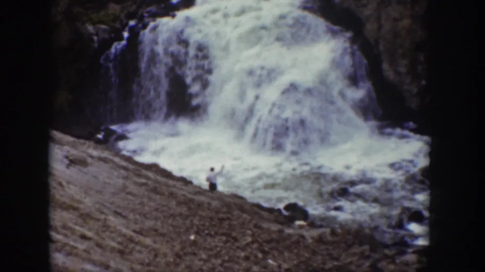 1939 Wyoming home-movie frame with a man standing on a rocky slope near a powerful waterfall.