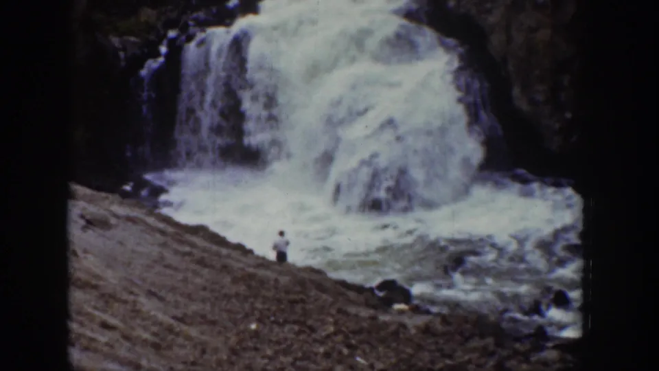 1939 Wyoming home-movie frame with a man near the base of a foaming waterfall among dark rocks.