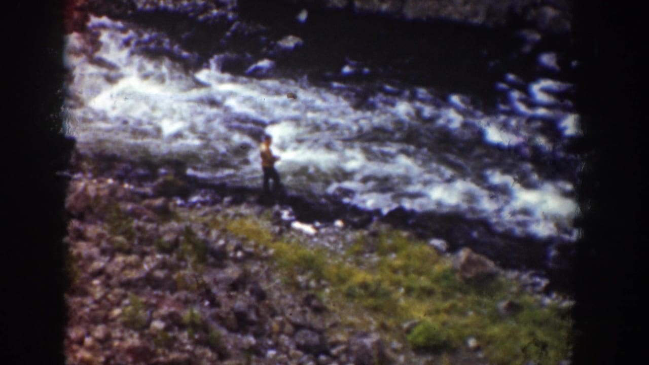 1939 Wyoming home-movie frame showing two people near a fast rocky river, one in a yellow shirt, with dark film edges visible.