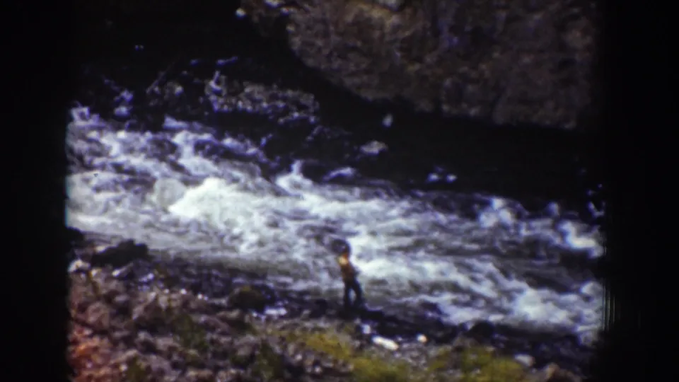 1939 Wyoming 8mm frame with a distant figure on the riverbank beside foaming rapids and dark rocks.