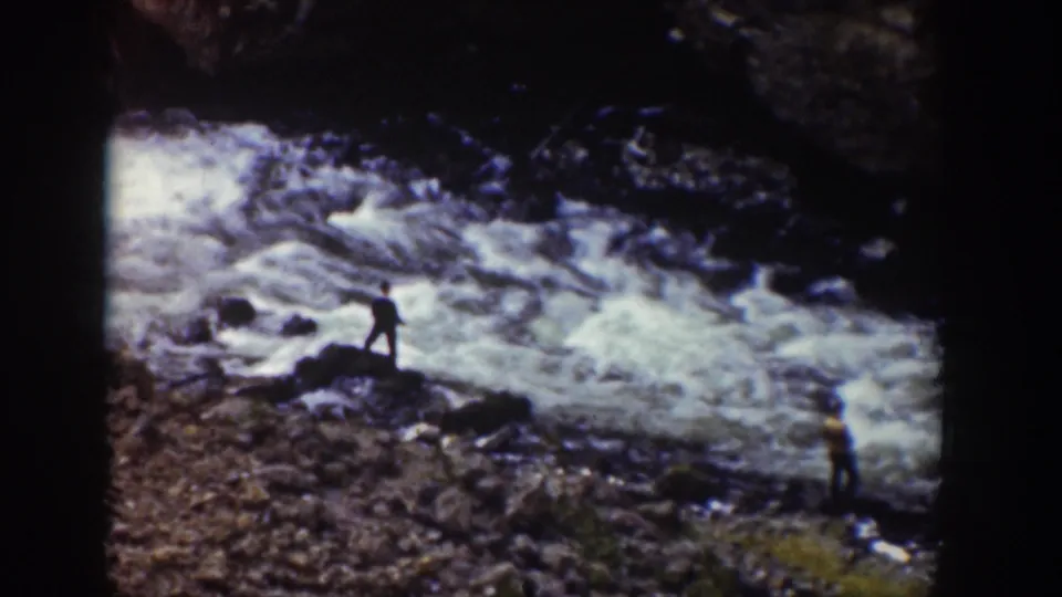 1939 Wyoming home-movie frame showing two distant people near a rapid river, with one figure in silhouette and another in a yellow shirt.