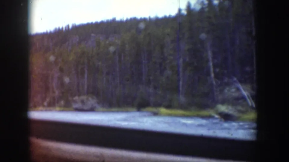 1939 Wyoming 8mm frame showing a wider view of the river, evergreen forest, rocky bank, and soft color-faded film texture.