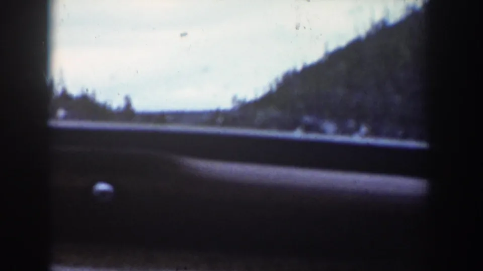 A blurred passenger view shows roadside water, a wooded slope, and distant valley forms over the dark edge of the car door.