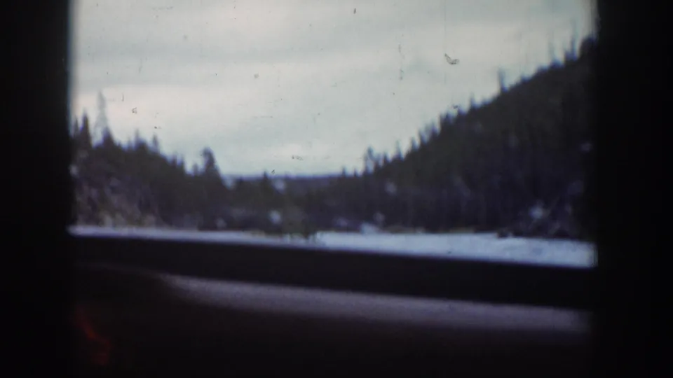 The car window frames a Wyoming valley view with water, dark wooded hillsides, and a pale overcast sky.