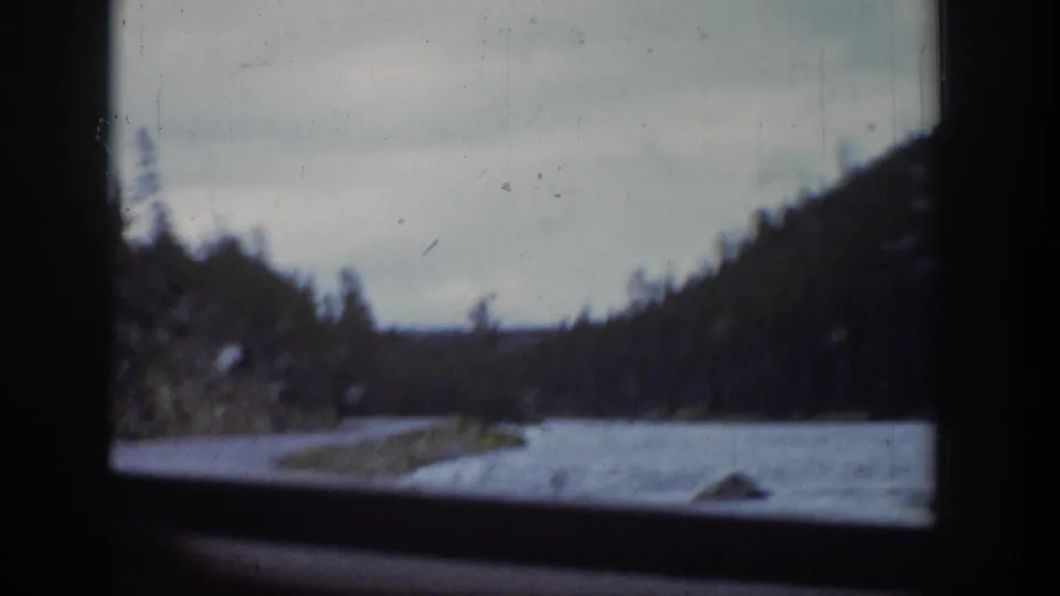 Through the car window, a Wyoming river and snow-lined bank appear below evergreen hills in soft 8mm focus.