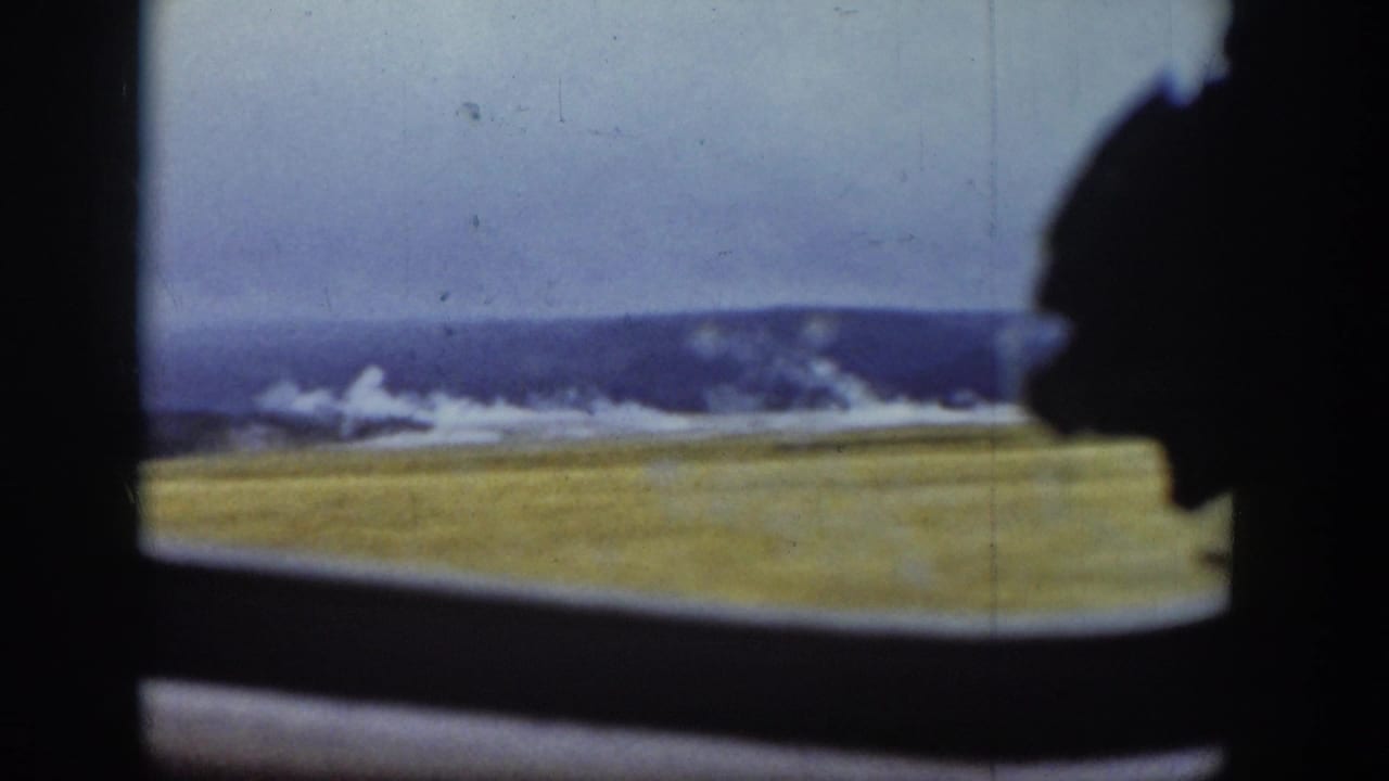 View through a dark vehicle window toward grassy plains, distant hills, and pale sky.