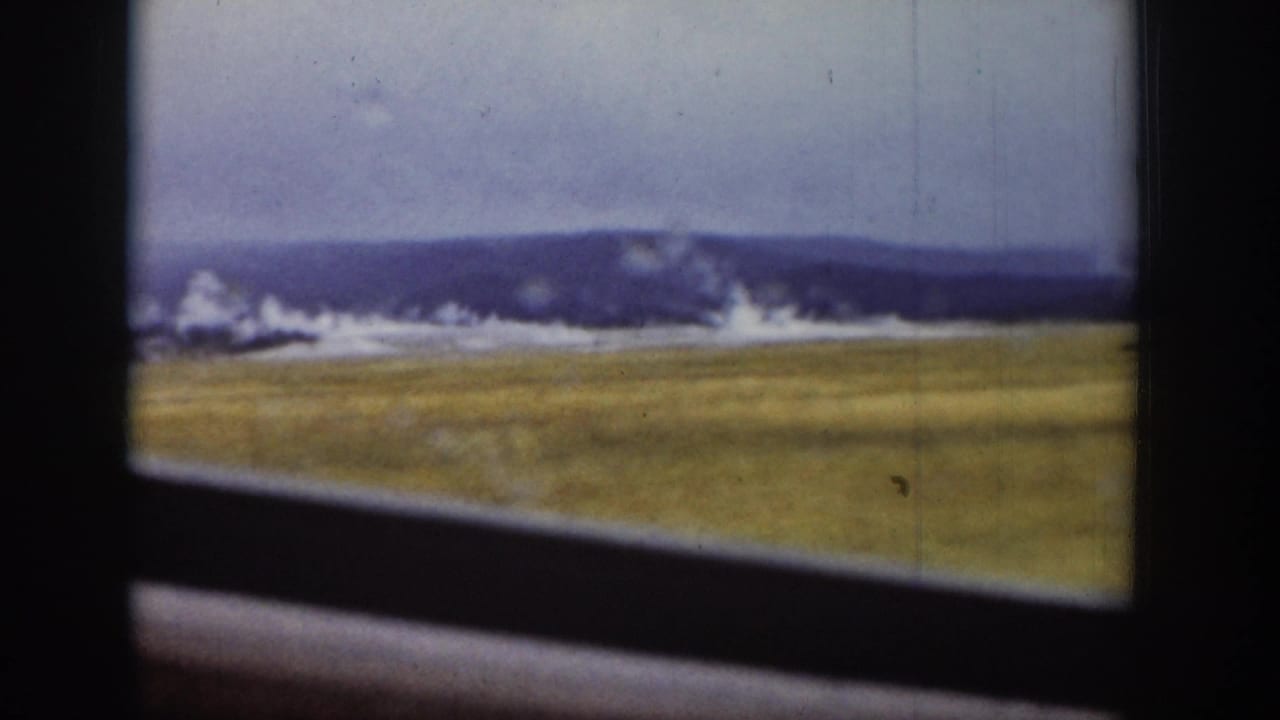 Distant geyser steam seen through a vehicle window with field and ridge beyond.