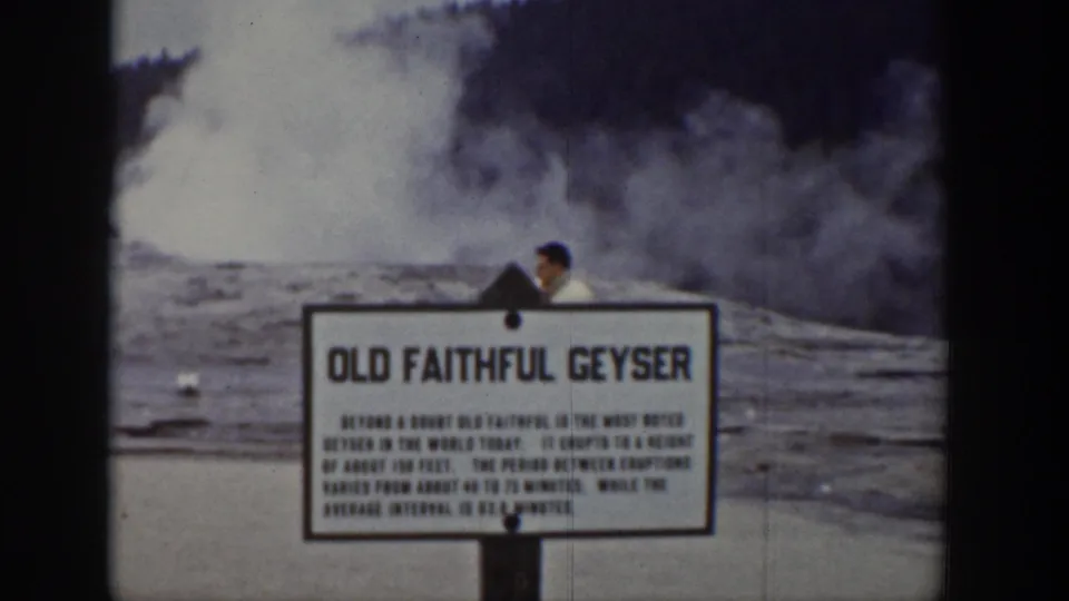 1939 Yellowstone home-movie frame showing the Old Faithful Geyser sign, steam in the background, and a visitor partly visible nearby.