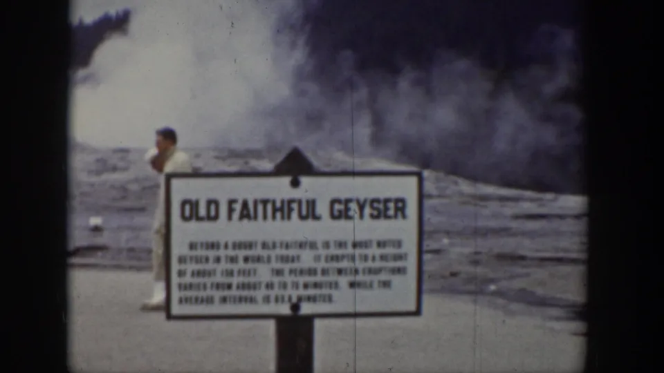 1939 Wyoming frame with the Old Faithful Geyser sign centered, a visitor behind it, and a broad steam cloud over the basin.