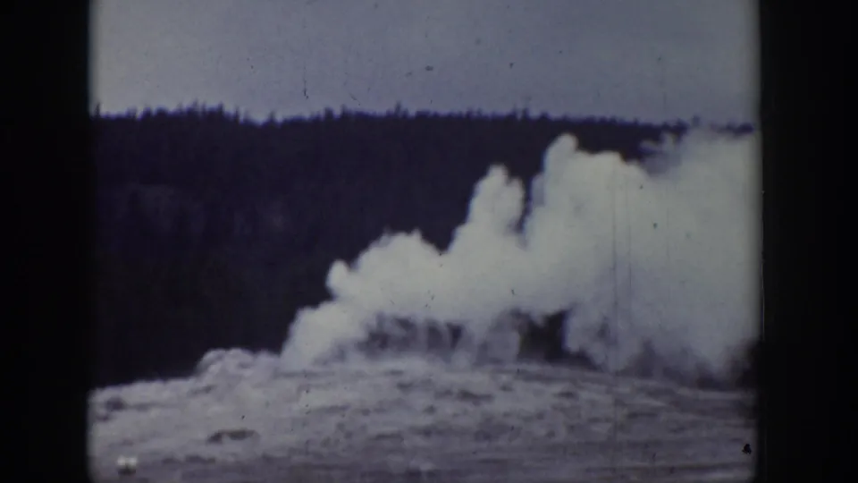 1939 Wyoming home-movie frame showing Old Faithful erupting, with white steam and water rising against a dark forested backdrop.