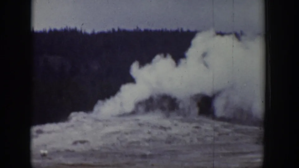 1939 Wyoming home-movie frame showing a white geyser plume rising near a dark conifer forest edge.