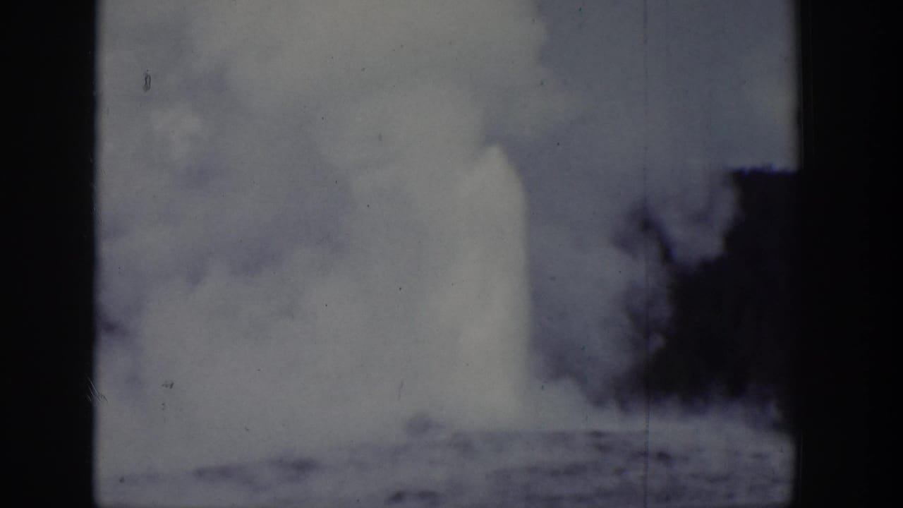 1939 Wyoming home-movie frame showing a distant geyser eruption with steam or water rising behind trees.