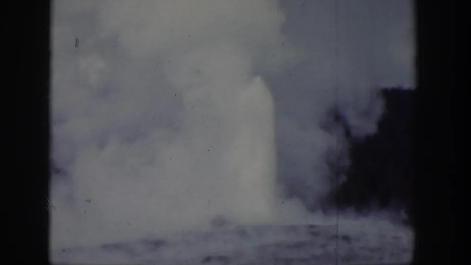 1939 Wyoming home-movie frame showing a distant geyser eruption with steam or water rising behind trees.