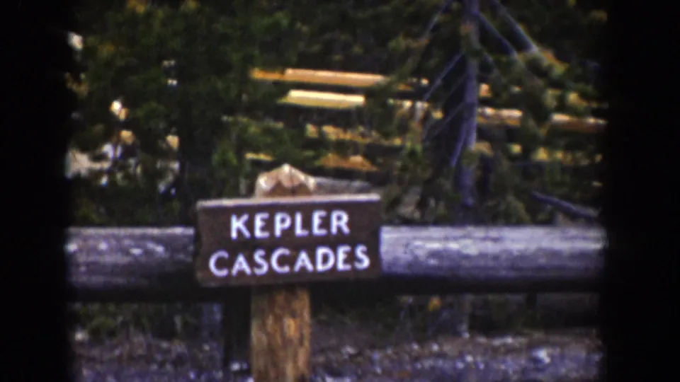 1939 Wyoming home-movie frame showing a rustic Kepler Cascades sign beside a log fence in evergreen forest.