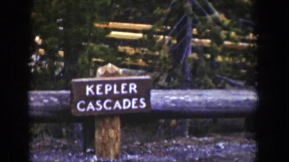 1939 Wyoming 8mm frame with a readable Kepler Cascades sign set against evergreen forest and a rustic log rail.