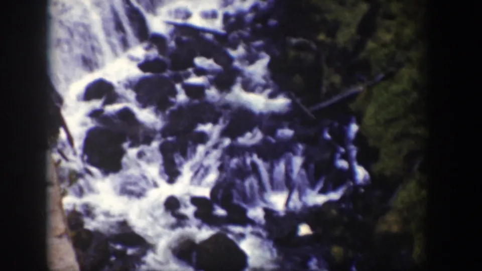 Close 8mm view of turbulent Wyoming stream water moving over dark rocks, with forest shadow and a strip of film edge visible.