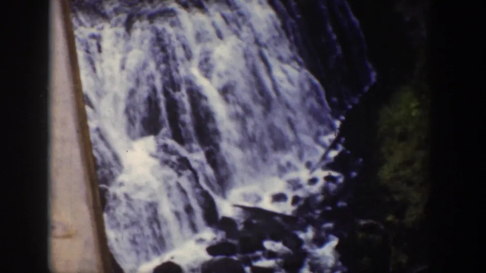Close 1939 Wyoming 8mm frame of a small waterfall spilling over dark rock, with wooded bank and film-edge shadow at the side.