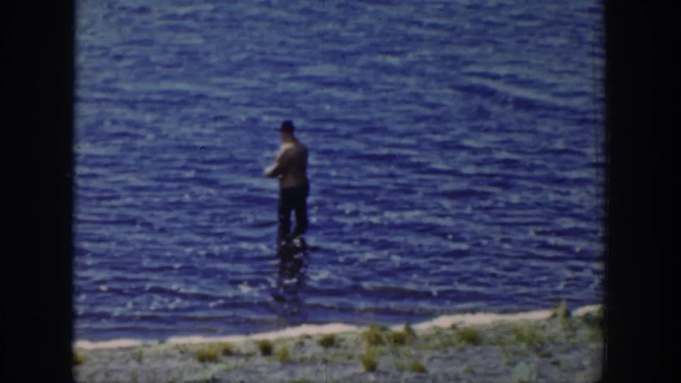 A distant fisherman stands in blue lake water, framed by soft 8mm texture and metadata-supported Wyoming location.