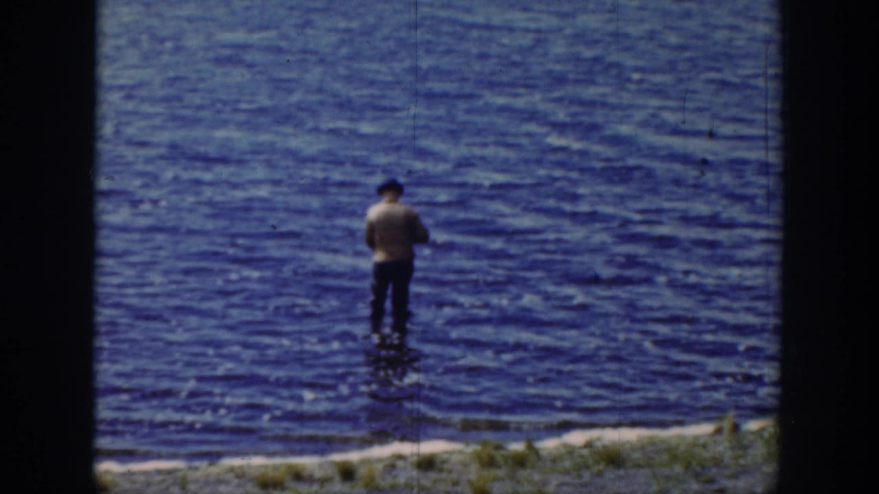 1939 Wyoming home-movie frame showing a man in a dark hat and light shirt fishing in shallow rippling water near grass.
