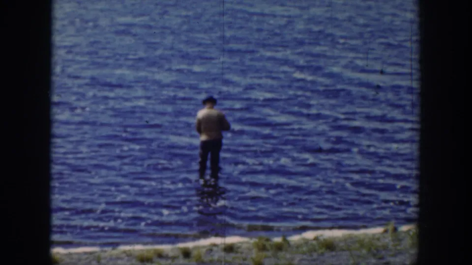 1939 Wyoming home-movie frame showing a man in a dark hat and light shirt fishing in shallow rippling water near grass.
