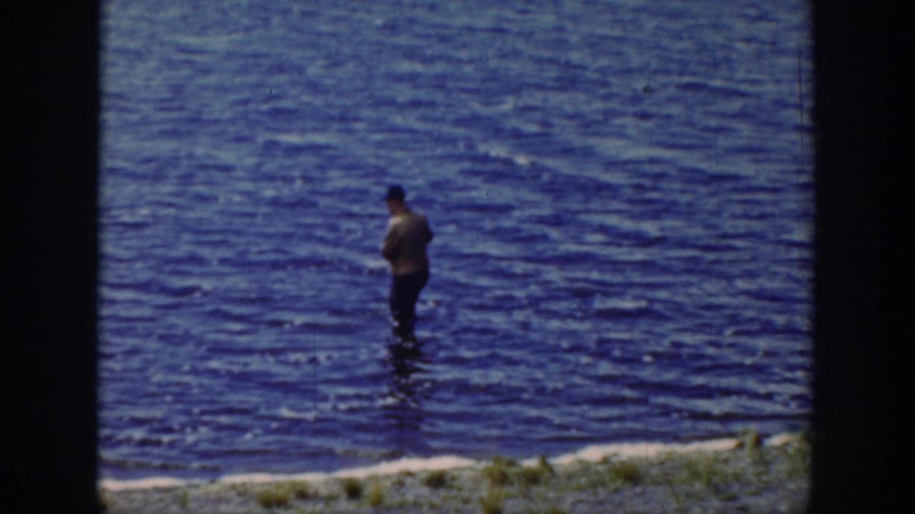 1939 Wyoming frame with a distant man holding a fishing rod in blue creek water beside a grassy edge.