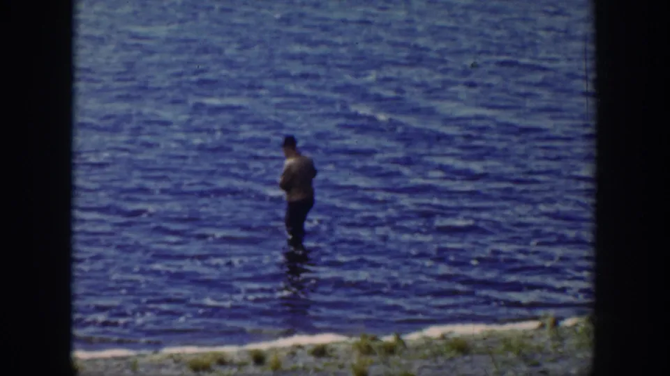 1939 Wyoming frame with a distant man holding a fishing rod in blue creek water beside a grassy edge.