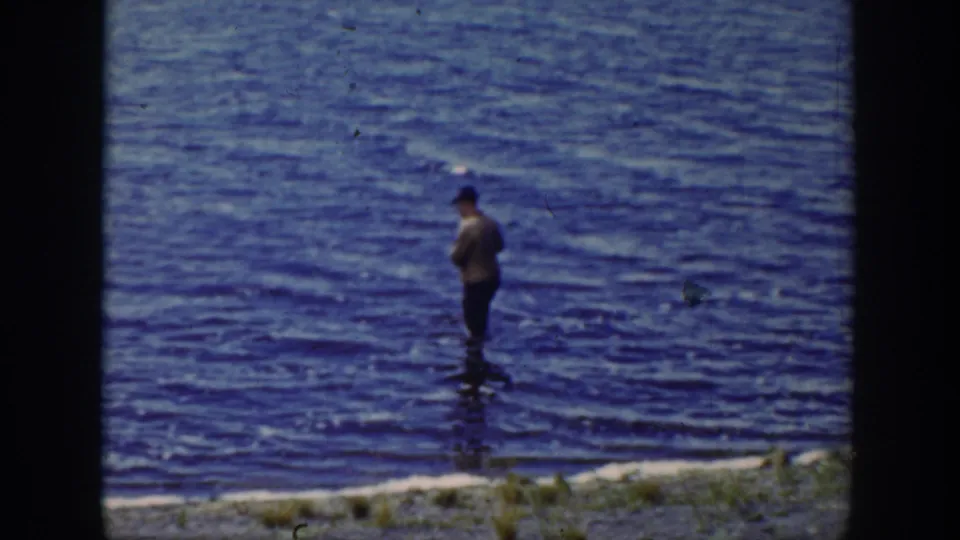 1939 Wyoming home-movie still of a man fishing in shallow creek water with grass visible along the bank.