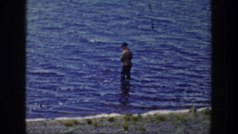 1939 Wyoming frame showing the fisherman in profile-like view amid rippling blue water and a grassy shoreline.
