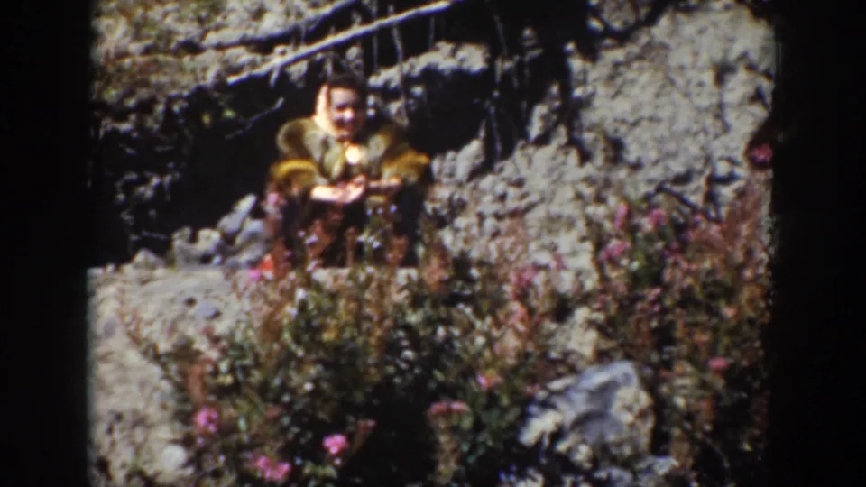 1939 Wyoming home-movie frame showing a woman in period clothing seated among rocks, brush, and pink wildflowers on a sunny hillside.