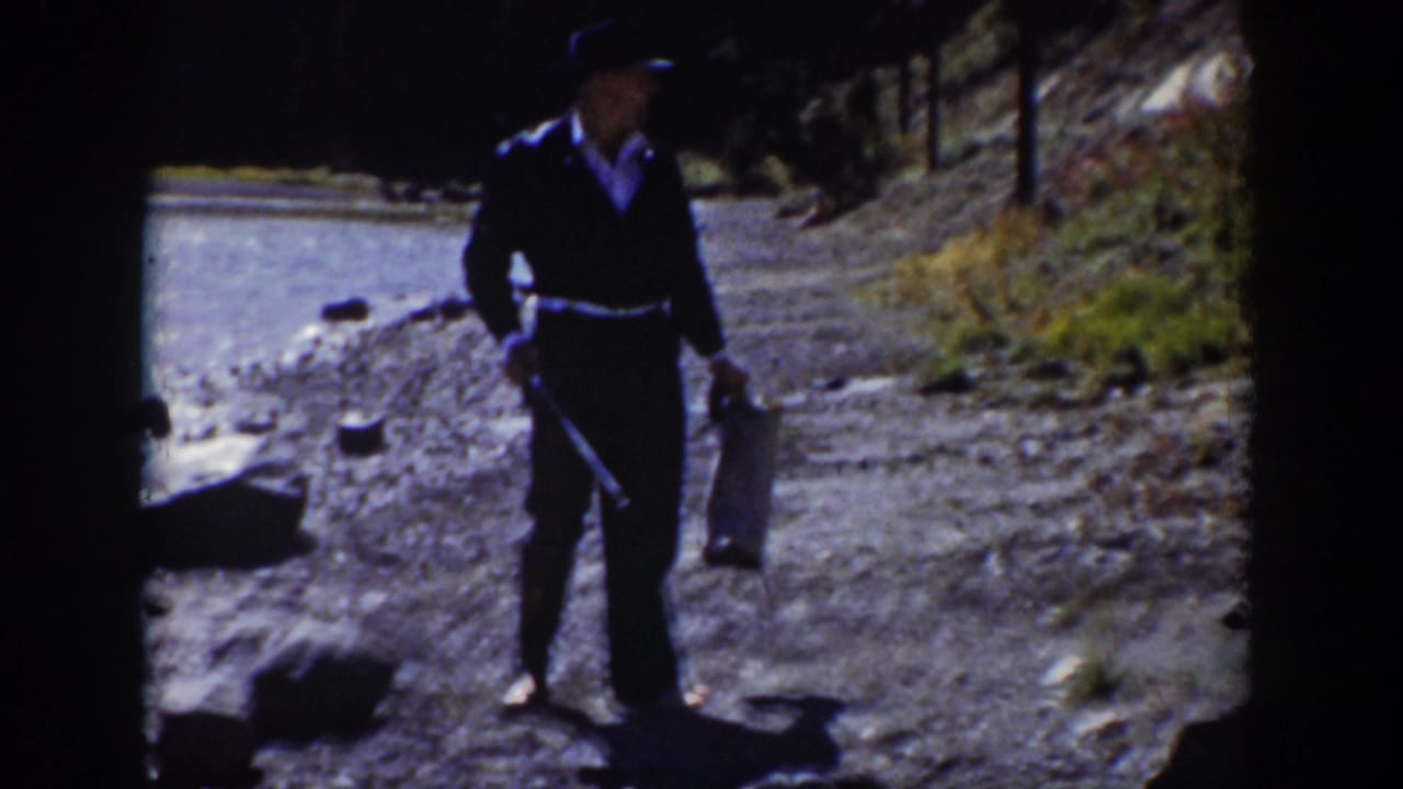 1939 Wyoming home-movie frame of a man in a dark suit and hat holding a bag and stick on a gravel riverbank.