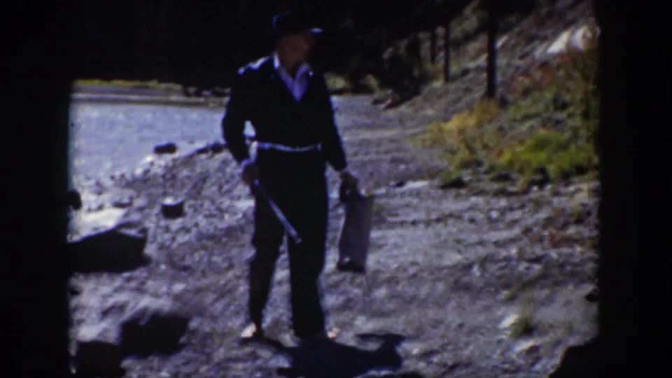 1939 Wyoming home-movie frame of a man in a dark suit and hat holding a bag and stick on a gravel riverbank.