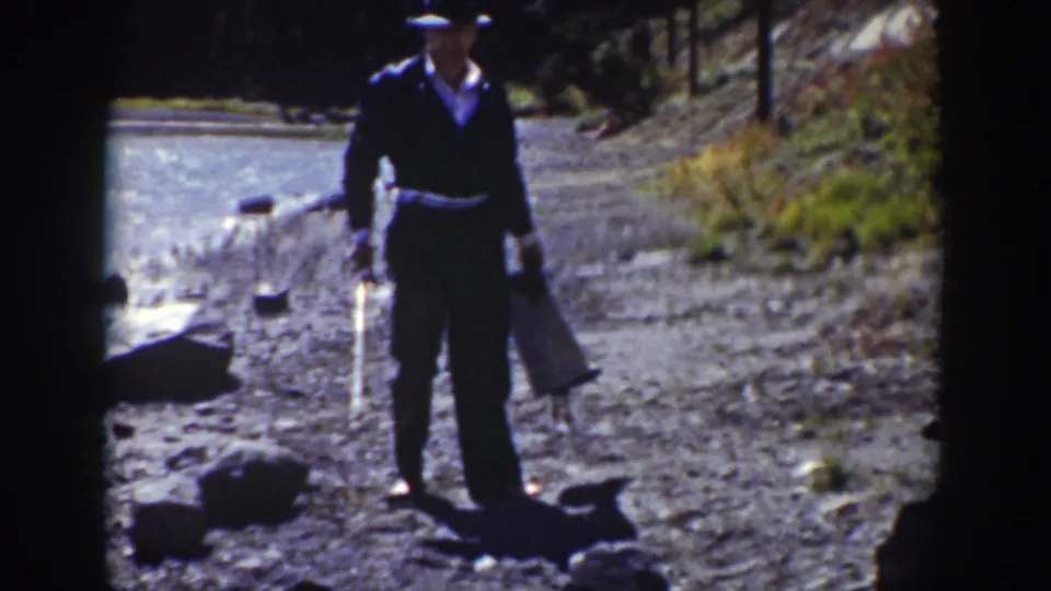 1939 Wyoming home-movie frame of a suited man facing the camera with a bag and stick on a sunlit riverbank.