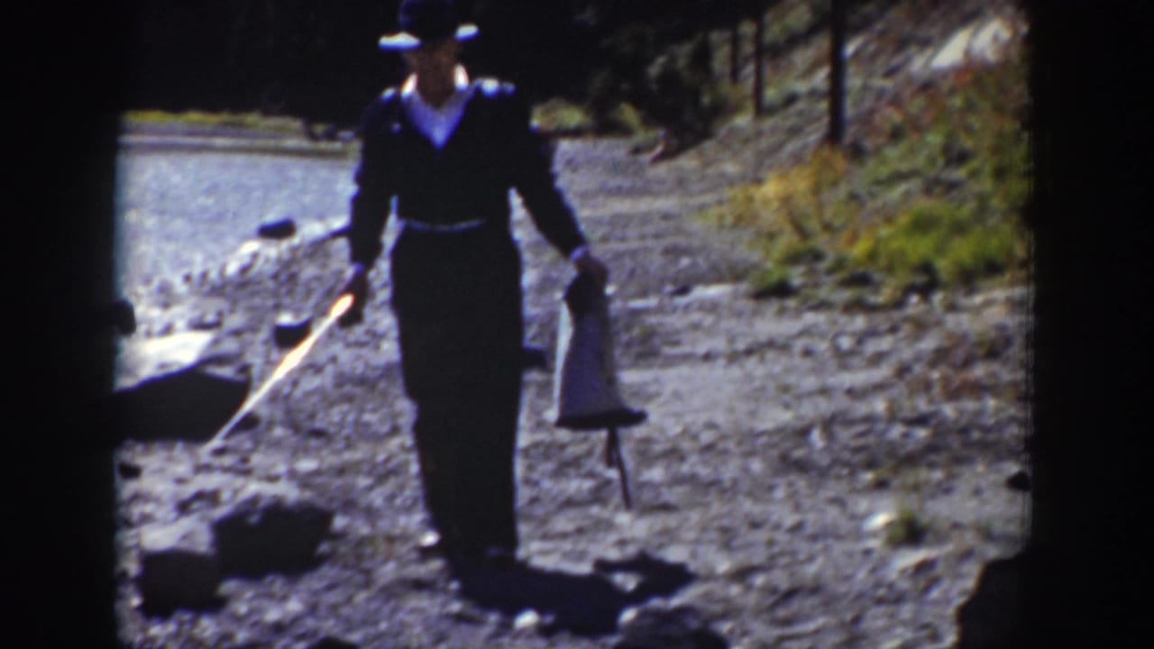 In metadata-supported 1939 Wyoming footage, a man in a dark suit and brimmed hat walks beside a rocky lake shore carrying fishing gear.