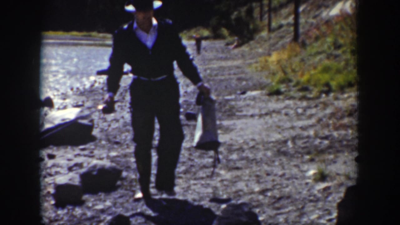 Man carrying fishing gear walks on a rocky shore near bright lake water.