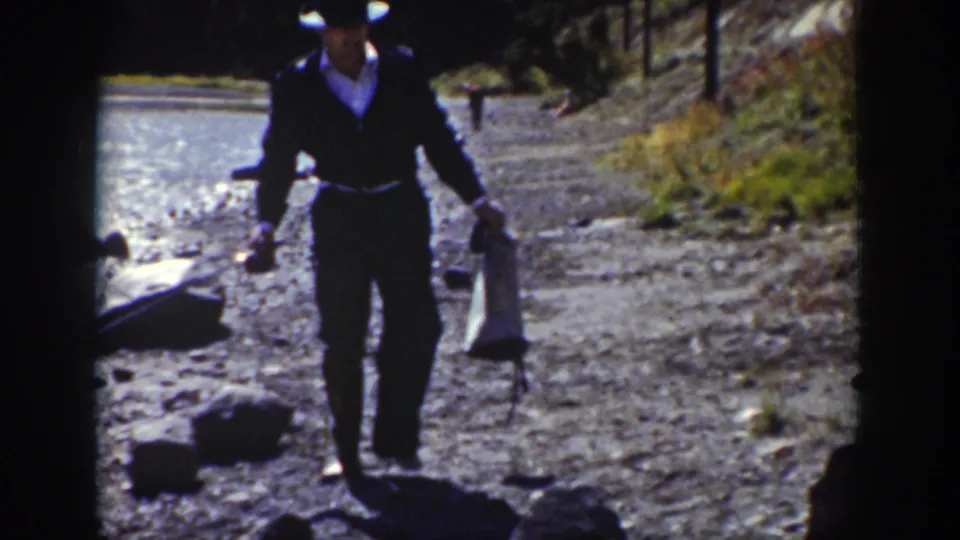 A suited man with a brimmed hat carries fishing gear along the rocky shore in metadata-supported 1939 Wyoming home-movie footage.