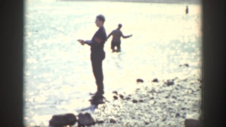 A man stands near the rocky waterline as another person wades in the bright lake, consistent with the Wyoming fishing scene metadata.